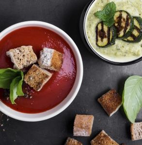 Bowl of red soup with croutons and garnish sitting next to additional croutons and another, smaller bowl with a green yogurt or dip with grilled cucumbers and garnish on top.