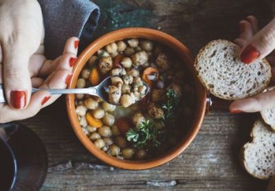 Woman's hands with red-painted nails holding a spoon scooping a chickpea-based soup or stew. Other hand is holding a piece of bread. The soup or stew is in a brown bowl, and it's sitting on a wooden table.