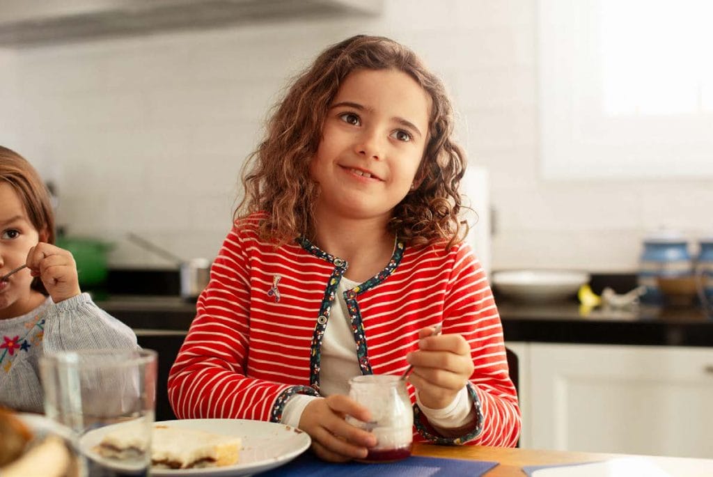 Child sitting at a kitchen eating a yogurt while listening something