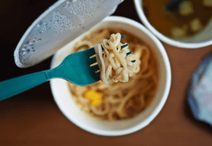Ramen in a paper cup with foil lid open, and a blue plastic fork picking up the ramen noodles.