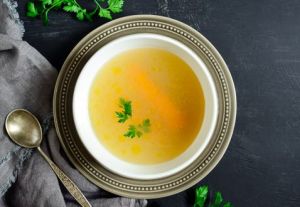 Clear-yellow soup or broth sitting in a white bowl and with garnish on top. White bowl is sitting on top of a silver charger with a silver spoon next to it. All on a black background or table with a leaf-like decoration running behind it.
