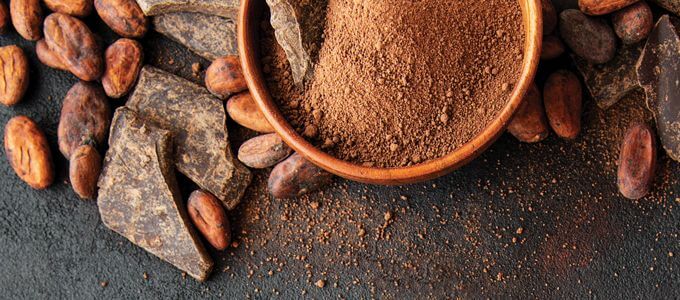 Bowl of cocoa powder in a bowl sitting on a table with some powder sprinkled around and cocoa beans and bars of chocolate around the bowl, as well