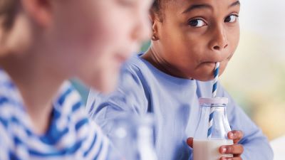Girl drinking milk out of a bottle with a straw, looking over at a friend who is closer to the camera but blurred out.