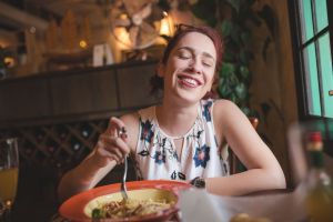 Woman with a floral blouse and bright red hair sitting at a table at a restaurant closing eyes and smiling, tilting her head. She's holding a fork that's spinning a pasta-like dish in a bowl. The setting is dimly lit and there is a fake plant, fan, decor and a wine rack in the background.