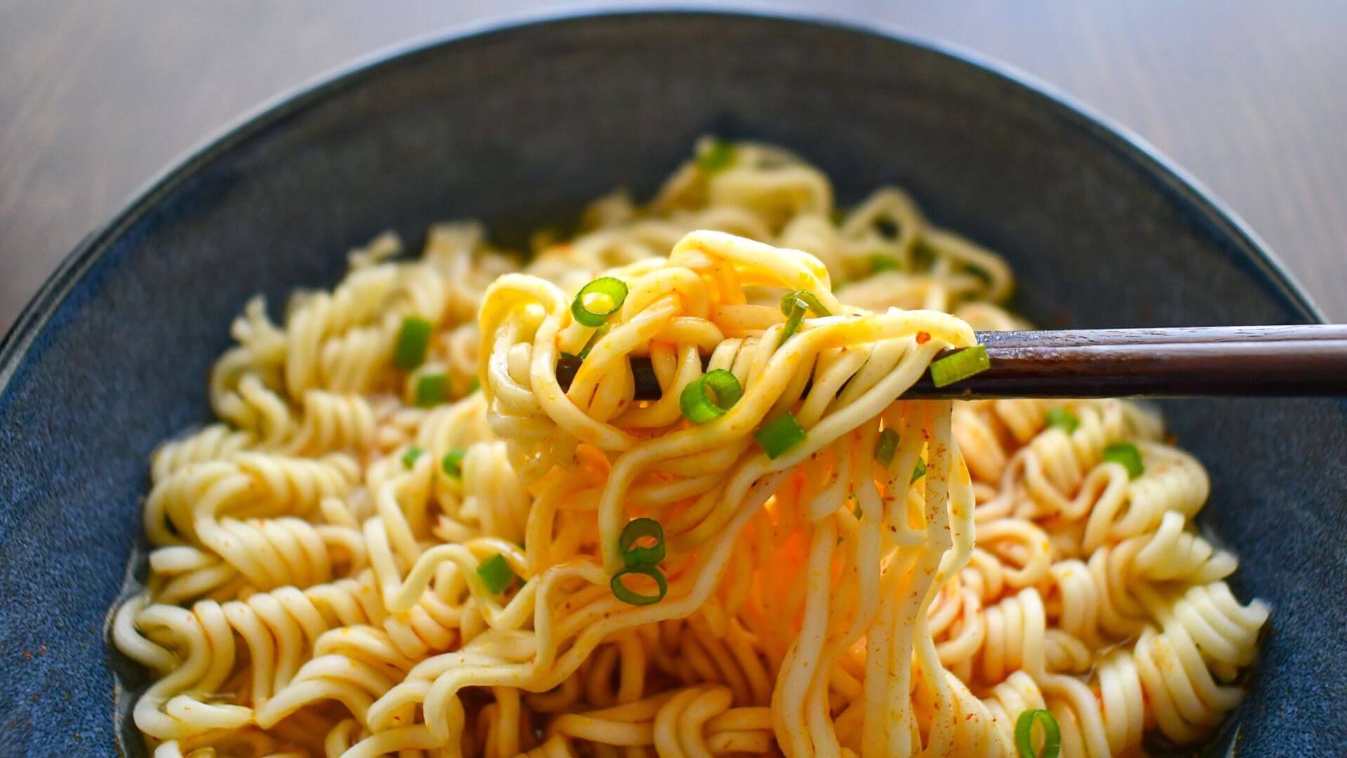 Ramen instant noodles in a bowl and set on a table. Noodles have seasoning and scallions on them, and a silver utensil is holding up some of the noodles.