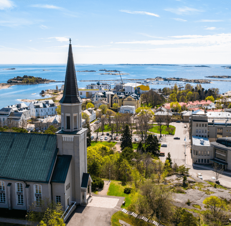 Aerial view of Hanko, Finland, with a church steeple in the foreground and a coastal landscape of buildings, trees, and islands under a clear sky.