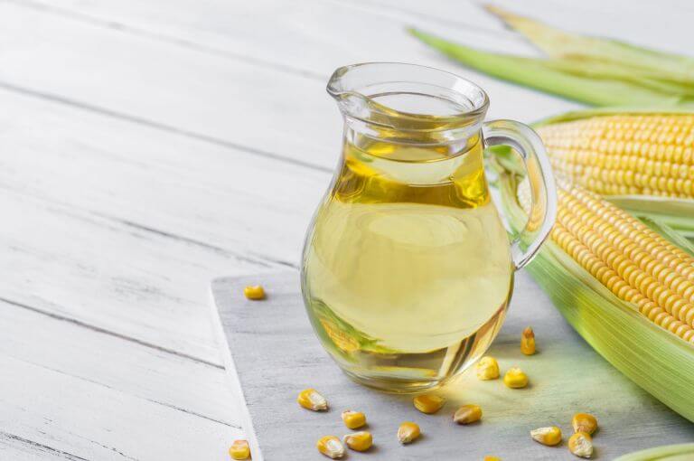 A pitcher filled with corn oil, accompanied by two corn husks full of corn on the side.