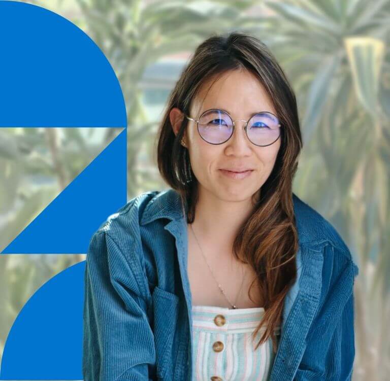 A woman with long brown hair wearing glasses, in a blue denim jacket and white top standing in front of a green background with a blue geometric design.