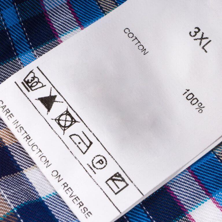A close-up of a clothing care label on a plaid cotton fabric, showing size 3XL and various laundry care symbols.
