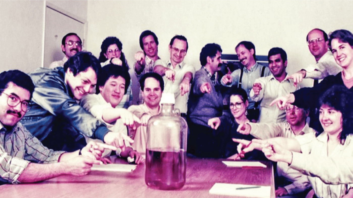 A group of people from Genencor gathered around a table, pointing toward a large glass container in the center.