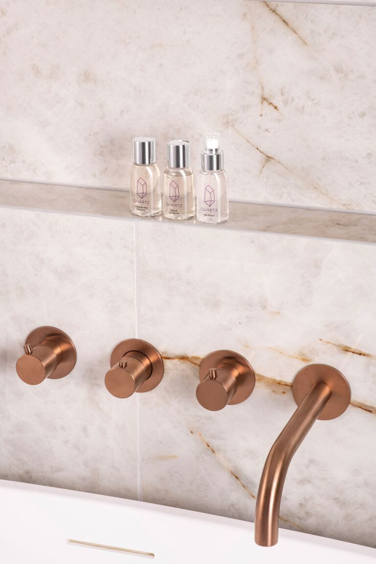 A bathroom wall with a marble finish, featuring three bronze faucet handles and a matching spout. Above the faucets, there is a shelf holding three small bottles of hair care products.