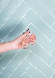 A hand holding two small bottles Quartz Shampoo and Conditioner against a light blue tiled background.