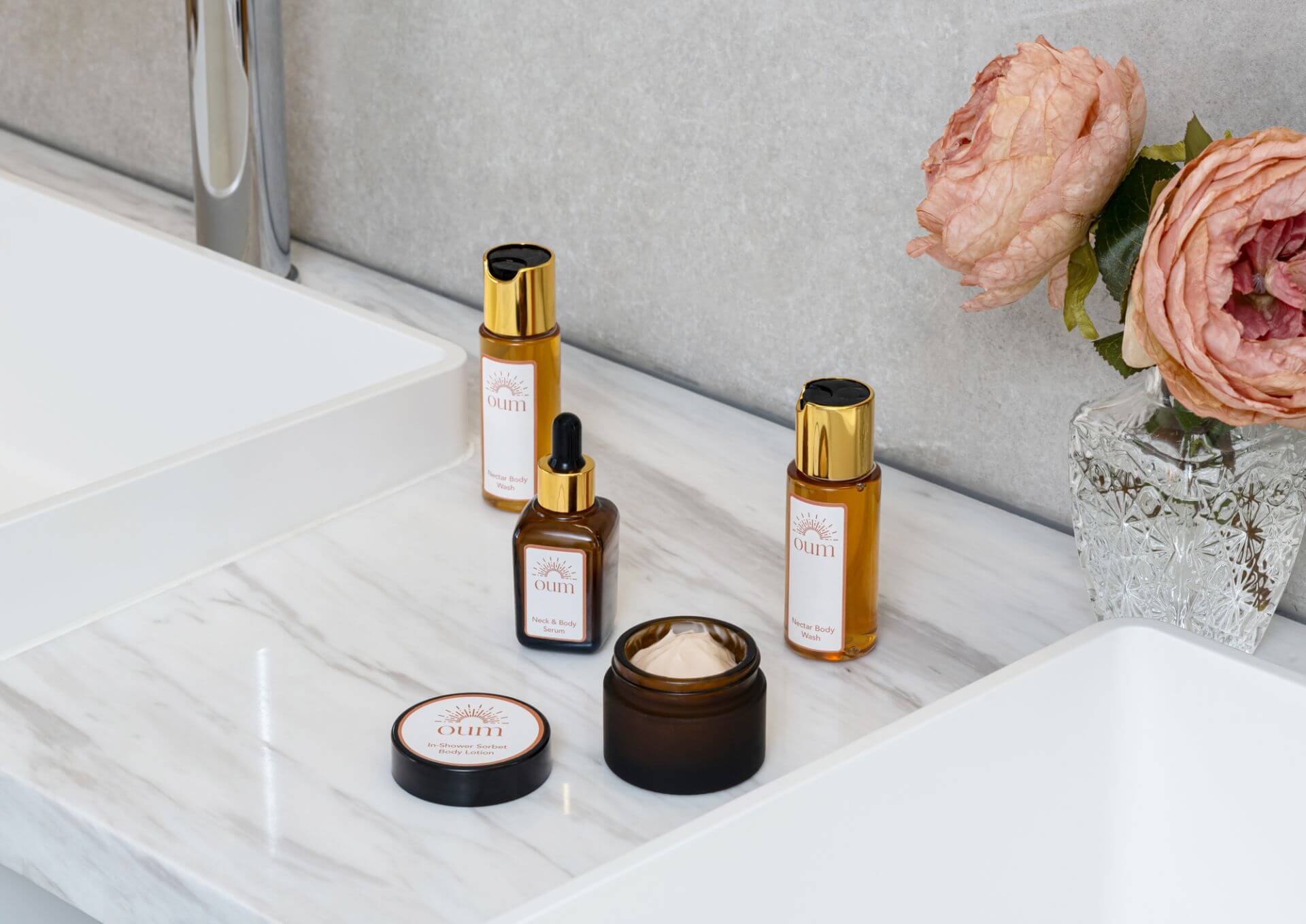 A bathroom countertop with bodycare products and a vase of pink flowers. The products include two pump bottles, a dropper bottle, and an open jar of cream.