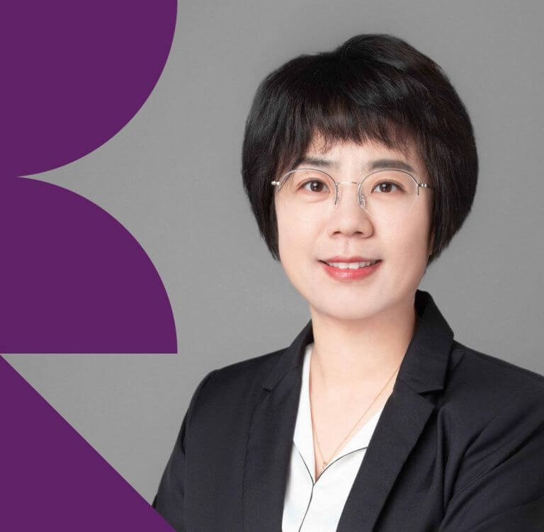 Woman with short bob cut black hair of east Asian heritage in a black blazer and white shirt with a gray background and purple geometric shapes.