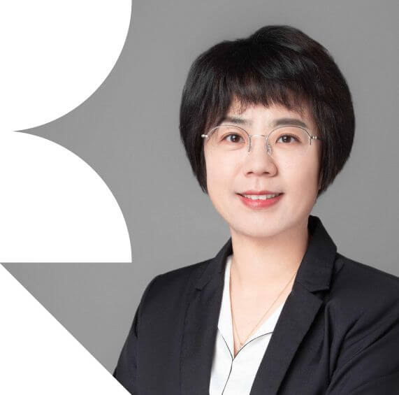 A woman, Lynn Meng, with short black hair, wearing a black blazer over a white shirt, stands against a gray background