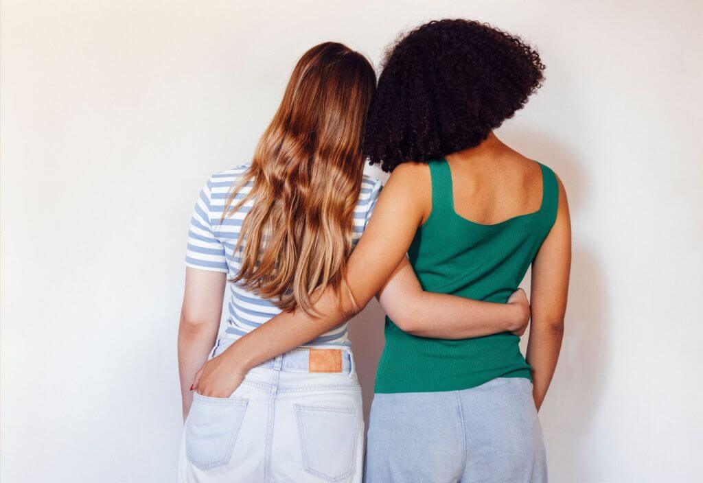 Two people standing side by side with their backs to the camera, arms around each other's waists. One person has long, straight brown hair and is wearing a striped shirt and white pants. The other person has short, curly black hair and is wearing a green sleeveless top and gray pants.
