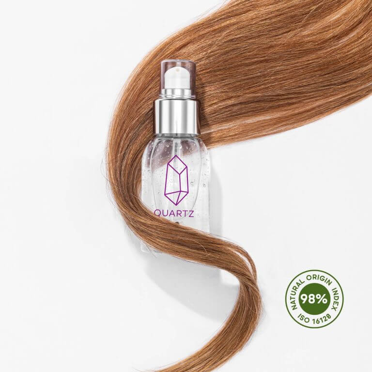 A bottle of Quartz hair serum with a pump dispenser is placed on a white background. A lock of long, straight, light brown hair is wrapped around the bottle. There is a green circular badge in the bottom right corner that reads 