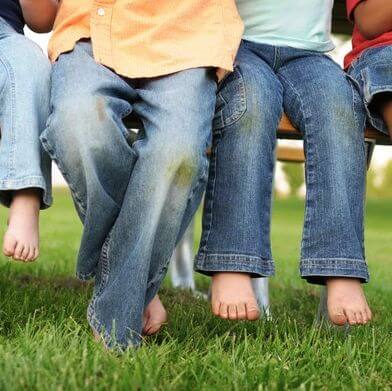 Close-up of young children on a bench, wearing blue jeans with grass stains, barefoot, with green grass beneath them.