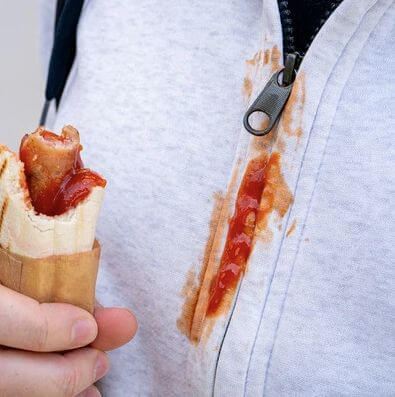 Young child holding a hot dog with ketchup, showing a visible ketchup stain on their shirt.