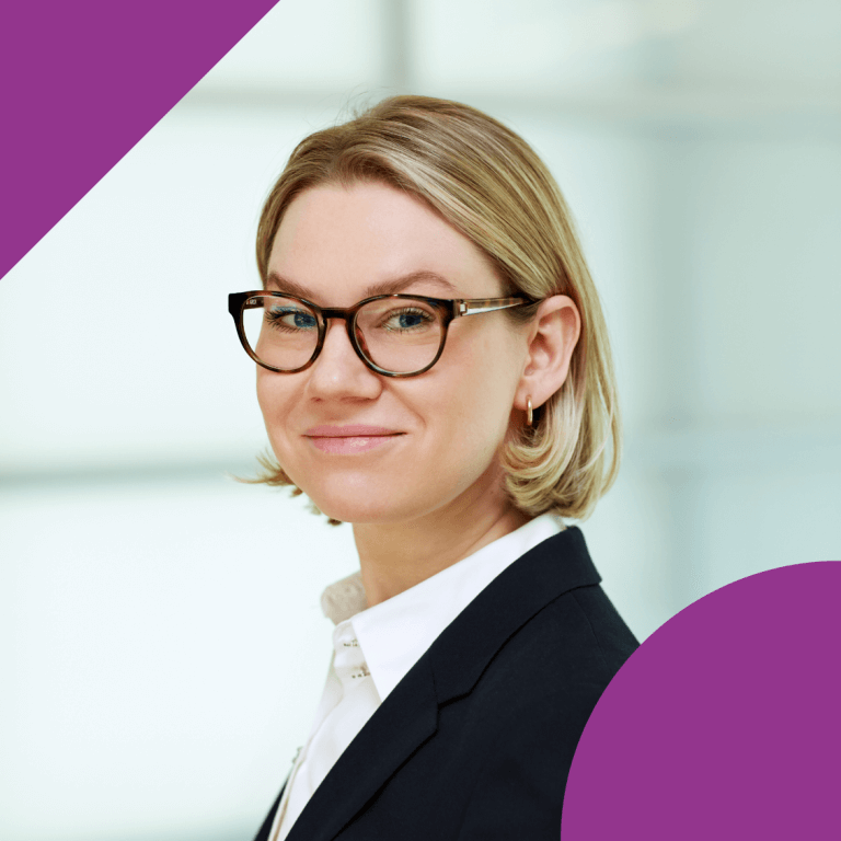 A woman in a black suit jacket and white shirt, with short blonde hair and glasses, stands in a softly lit indoor setting. Their face is obscured for privacy. Purple graphic shapes accent the image.