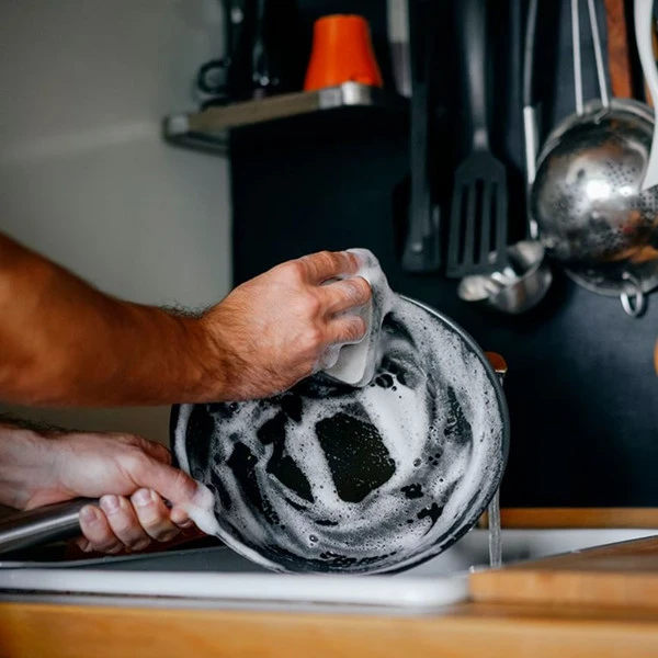 Close-up of hands washing a soapy black pan in a kitchen sink.