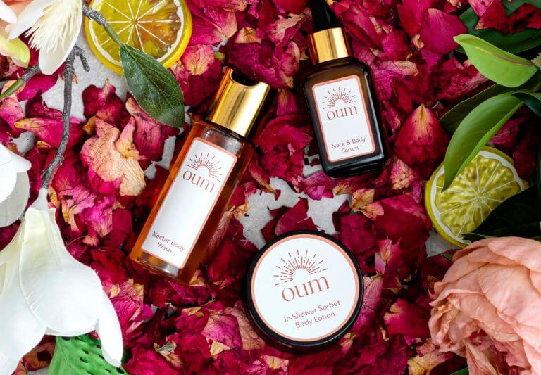 A flat lay image of bodycare products surrounded by pink rose petals, green leaves, and slices of lemon. The products include a bottle of 