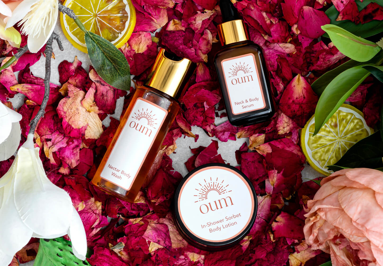 A flat lay image of bodycare products surrounded by pink rose petals, green leaves, and slices of lemon. The products include a bottle of 