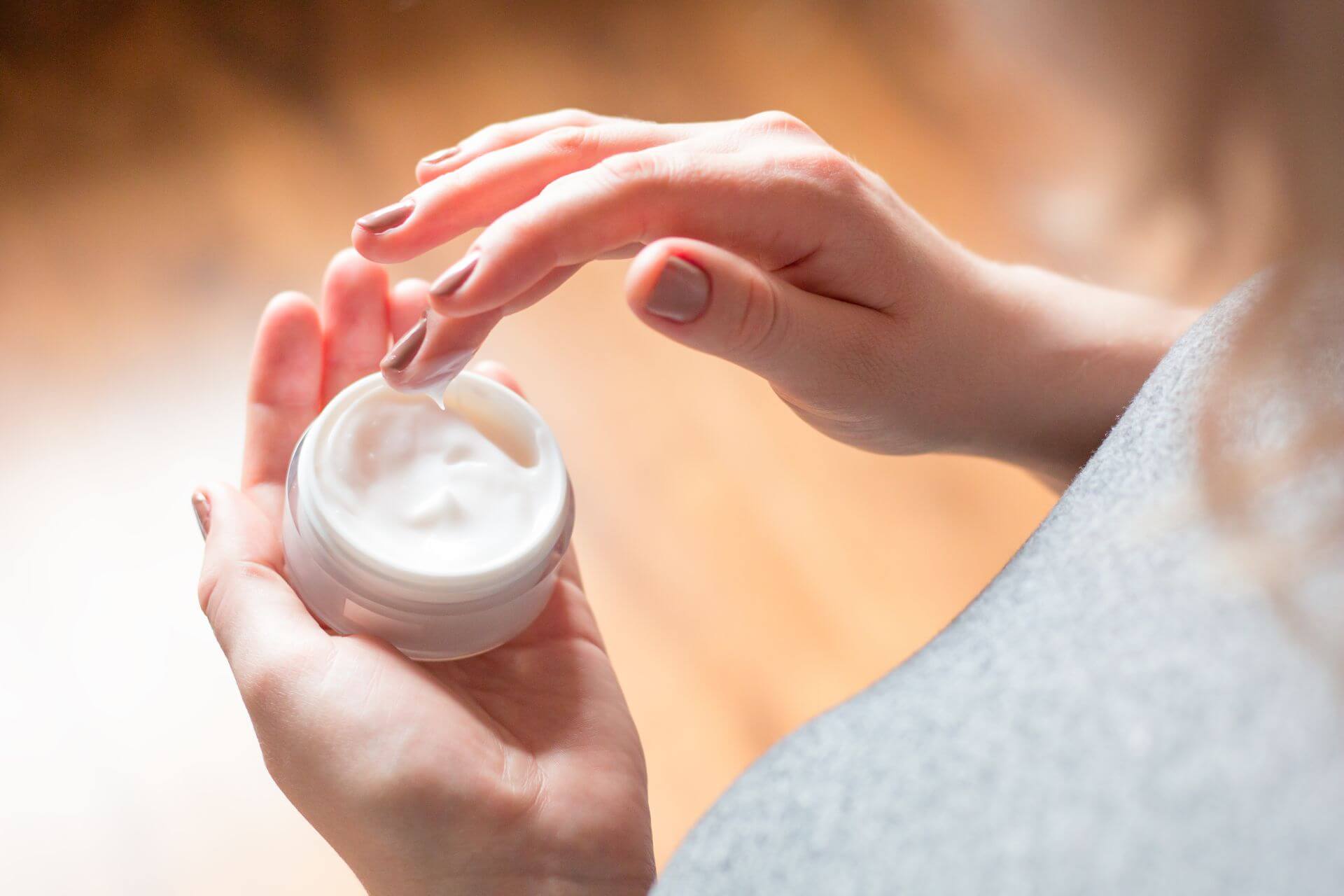 A person is dipping a finger into a small white jar of cream formulation, holding the jar with the other hand.