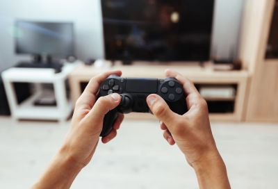 Hands holding a black video game controller in front of a television screen and entertainment center.