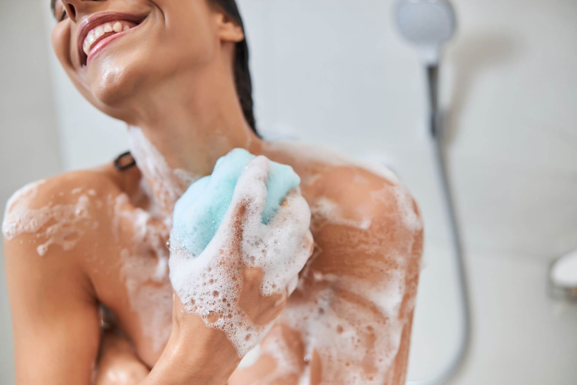A person in a shower, covered in soap suds, holding a blue sponge and smiling.