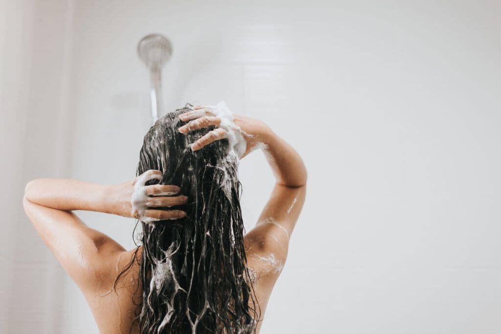 A person washing their hair in the shower, with their back to the camera. The person's hands are covered in shampoo lather as they scrub their wet hair.