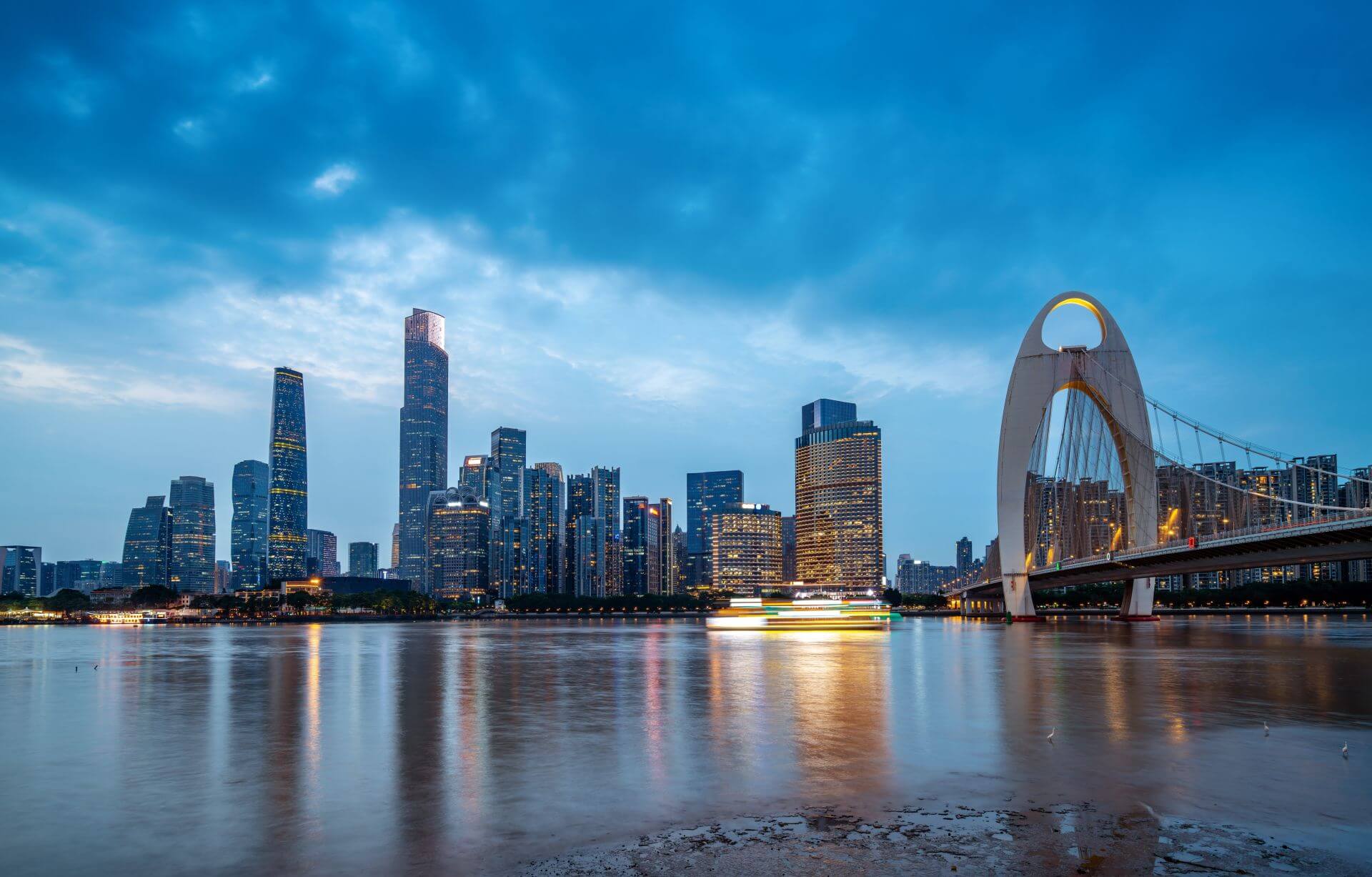 A panoramic view of a modern Chinese city skyline at dusk, with illuminated skyscrapers and a prominent bridge reflecting over a calm river (shutterstock_2474396631)