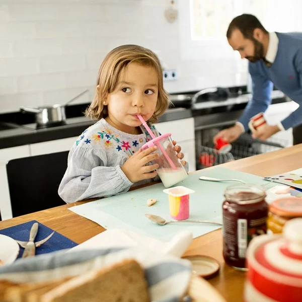 A child drinking from a pink sippy cup at a kitchen table while an adult loads a dishwasher in the background.