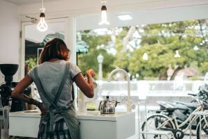 Woman in coffeeshop making and pouring coffee, not facing camera. Coffeeshop is bright and white, with a big window showing bicycles and greenery outdoors