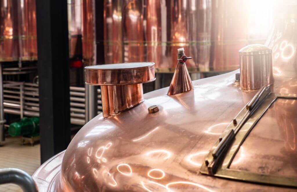 Close-up image of a copper brewing vat with various fittings and fixtures, reflecting light in a brewery setting