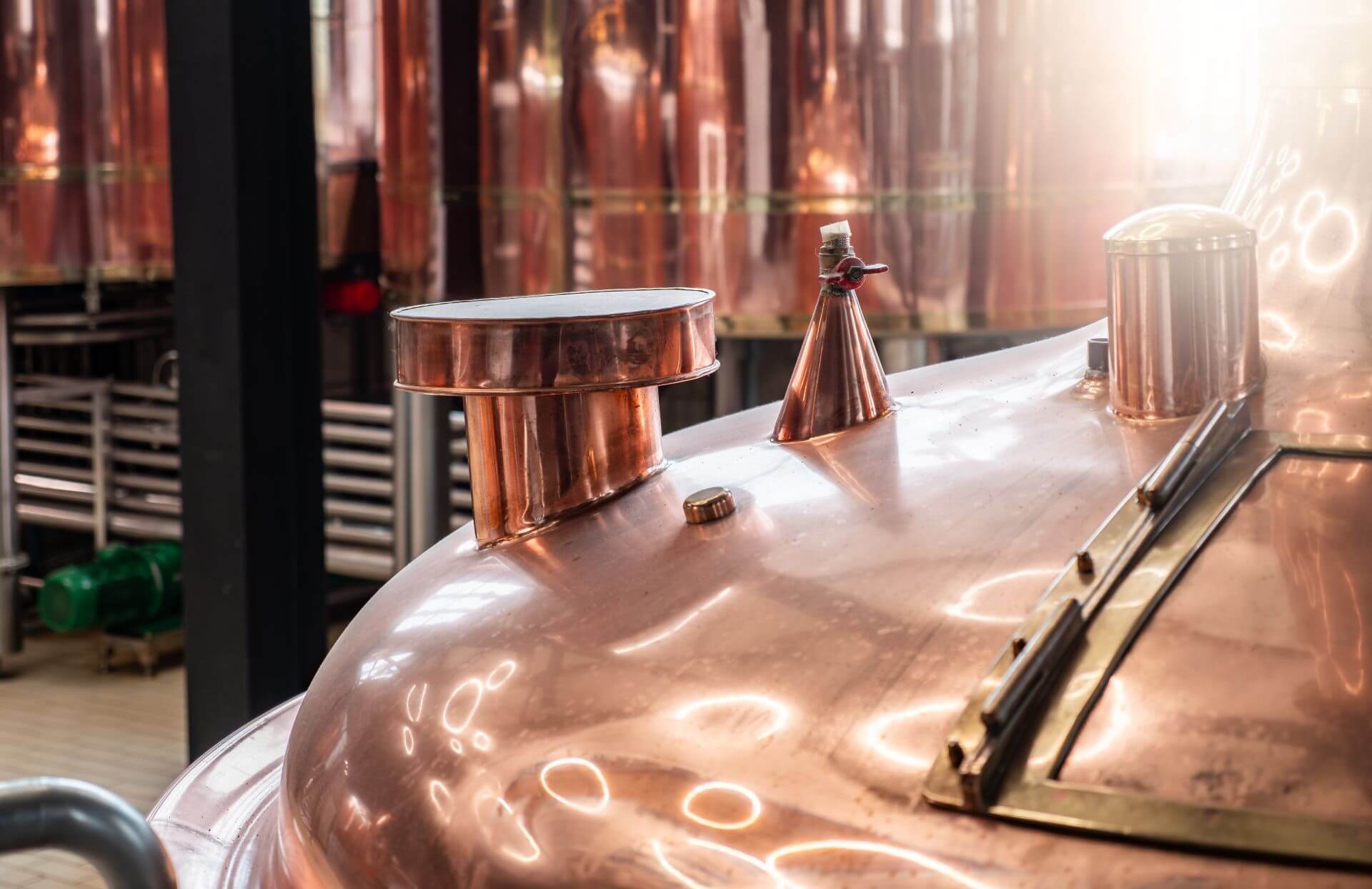 Close-up image of a copper brewing vat with various fittings and fixtures, reflecting light in a brewery setting