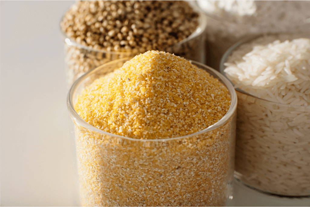 Close-up image of four glass containers filled with different types of grains and cereals: yellow cornmeal in the front, millet on the left, white rice on the right, and another type of grain partially visible in the background