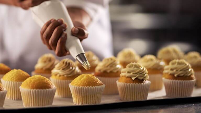 A row of multiple cupcakes with vanilla frosting being applied to each cupcake.