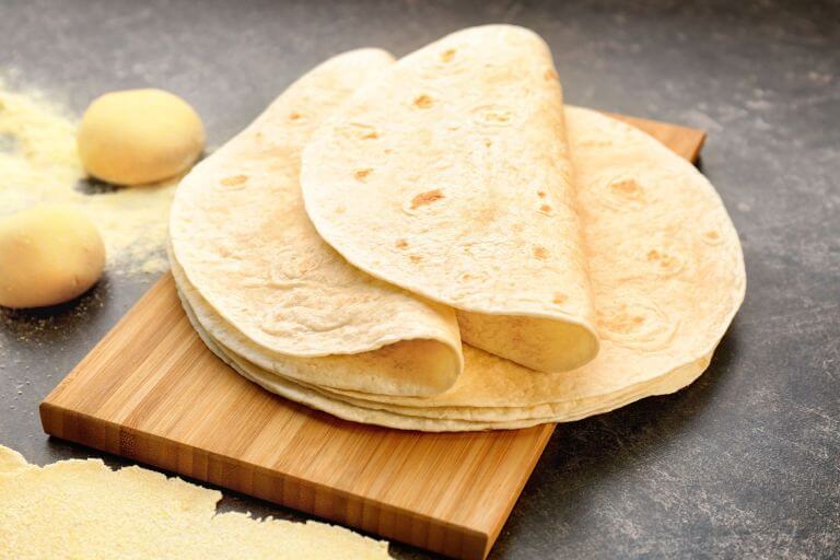 Flour tortillas on a wooden cutting board with dough balls and scattered flour in the background. POWERFLEX® enzymes maintain softness and moistness