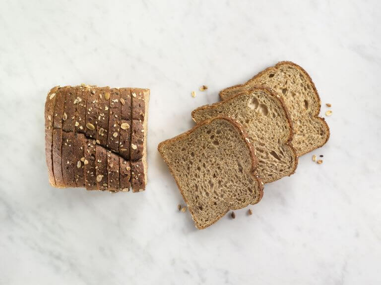 A loaf of sliced multigrain bread on a marble surface with several slices separated and laid out to the right. POWERFRESH® enzymes for baked goods freshness.