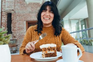 Woman with dark hair and yellow turtleneck eating cake in a cafe and drinking a latte-style beverage, smiling.