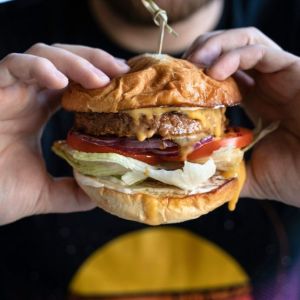 Close-up of hands holding a juicy burger with melted cheese, lettuce, tomato, and a toothpick on top.