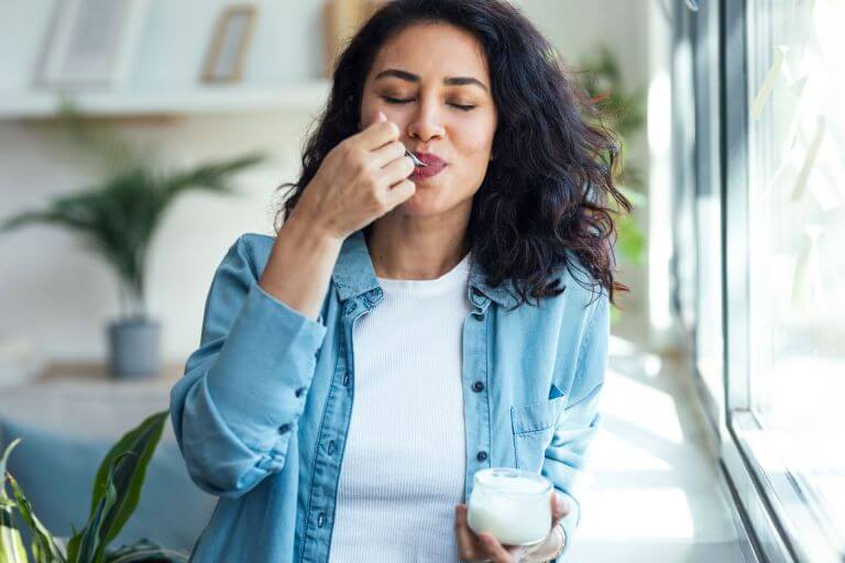 Person holding a yogurt cup and spoon, sitting by a window enjoying the texture and viscosity of a yogurt.