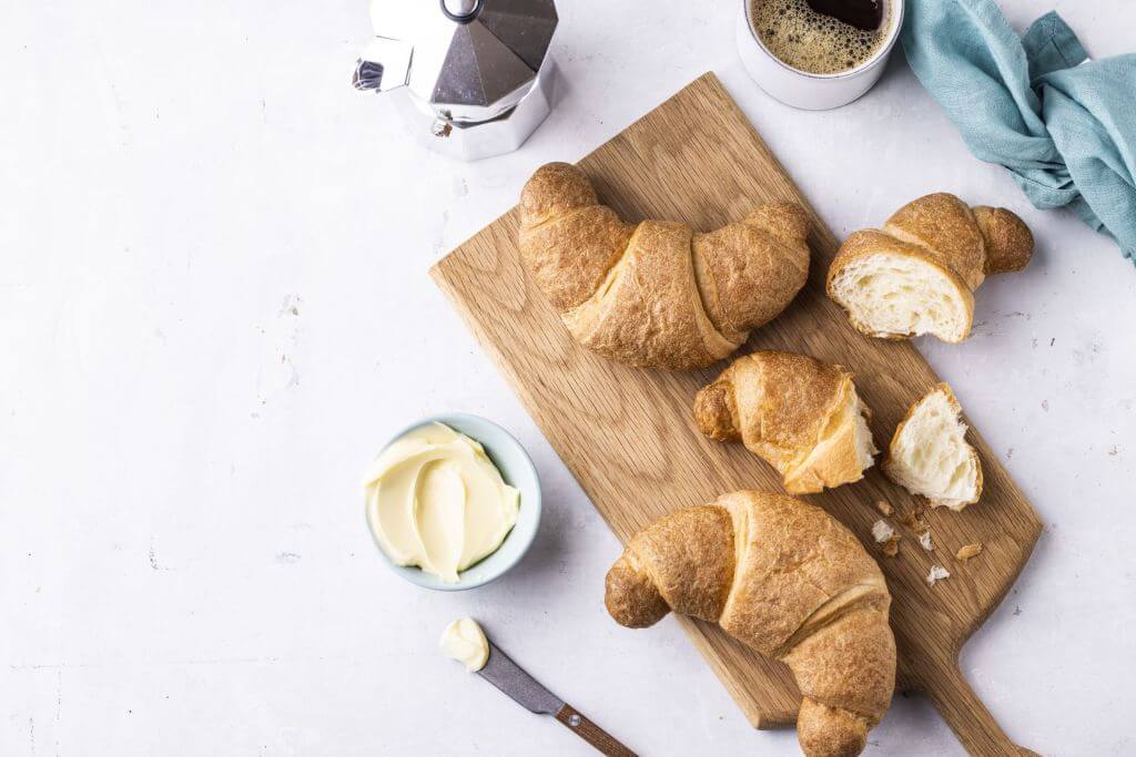 Wooden cutting board with three whole croissants and one partially sliced croissant, accompanied by a bowl of butter, a knife with butter on its blade, a cup of coffee