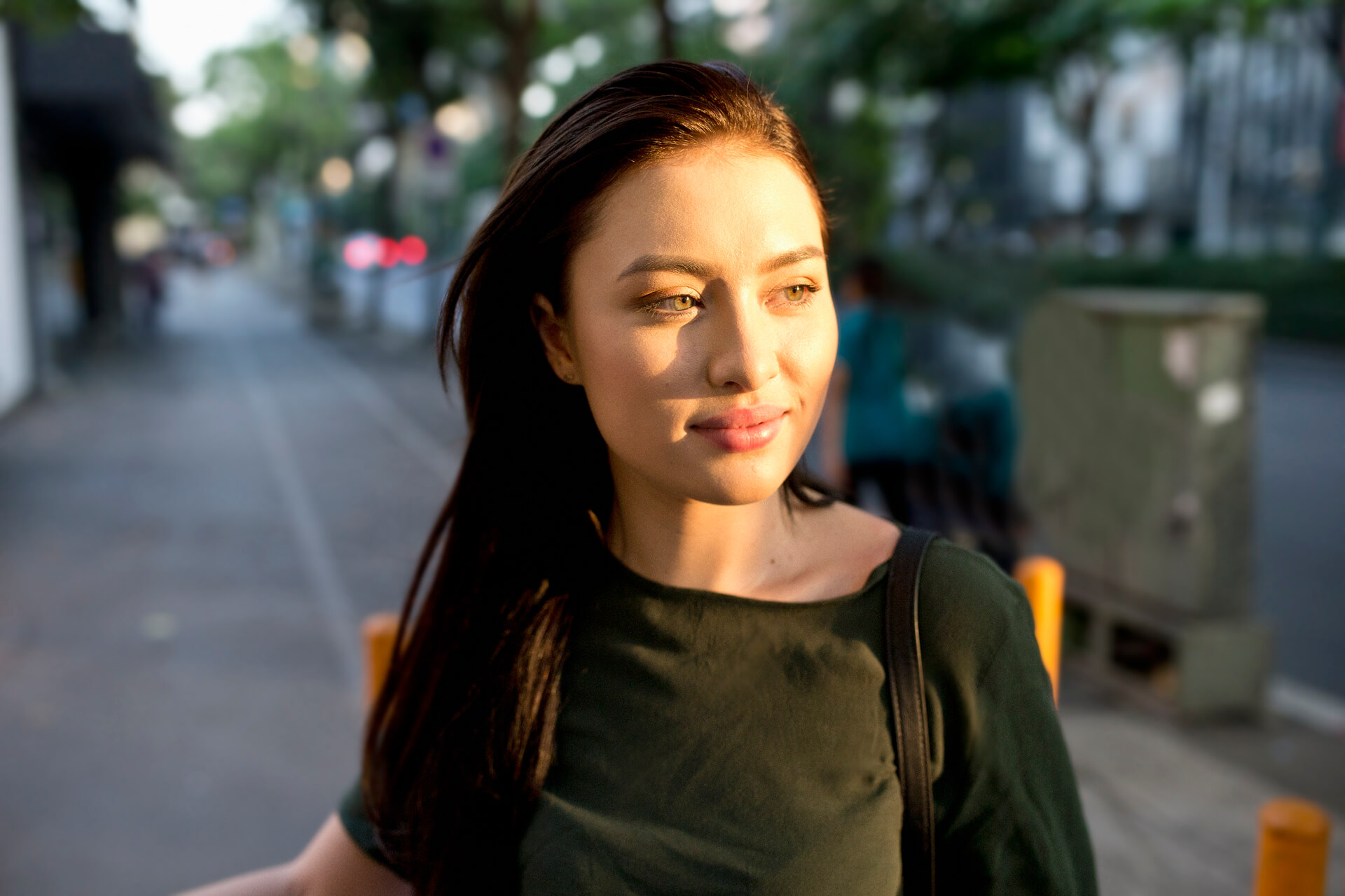 A person with long dark hair wearing a dark green shirt is standing on a sidewalk in an urban area.