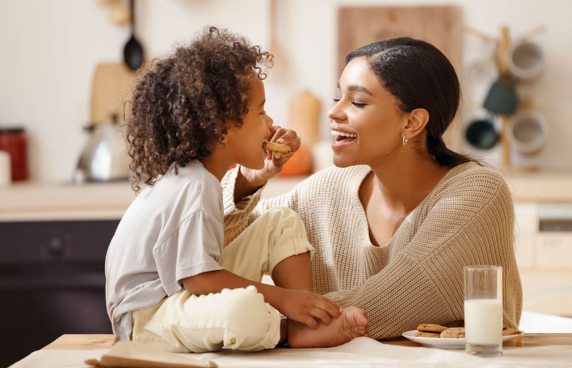 Mom smiling and looking at her child, feeding him a chocolate chip cookie. A plate of cookies and a glass of milk is on the table and they're in a kitchen setting