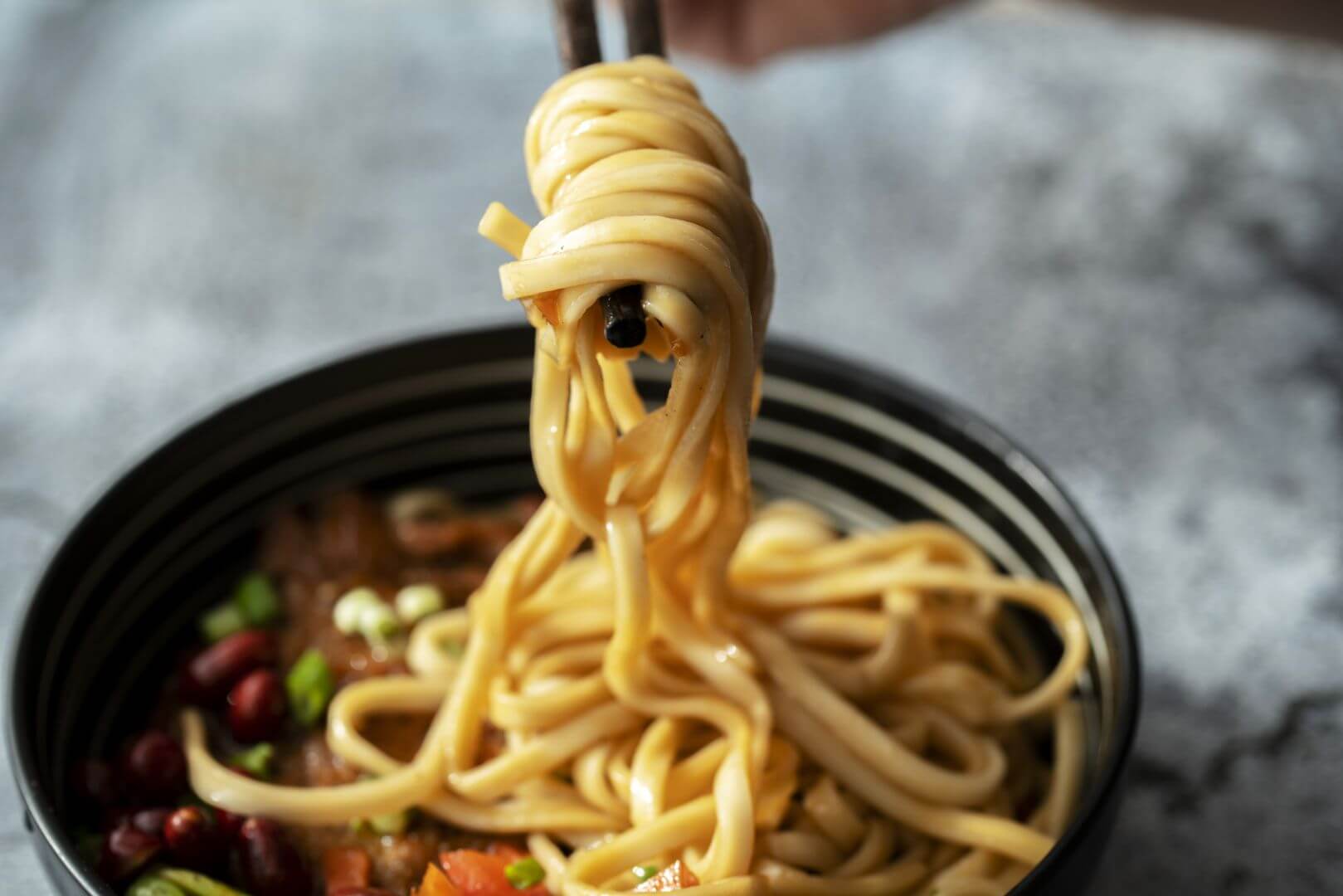 Chopsticks holding up and twirling noodles from a bowl of noodles, beans, vegetables and sauce