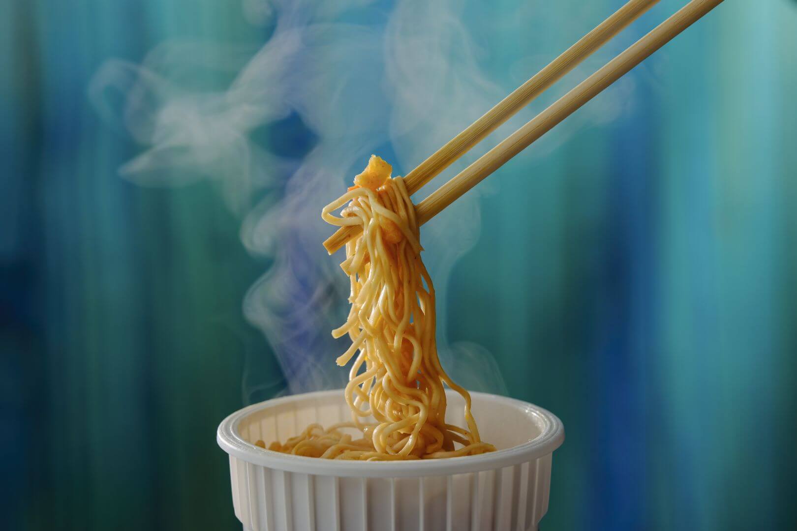 Chopsticks holding up noodles, out of a cup, with steam rising and a blue wall in background