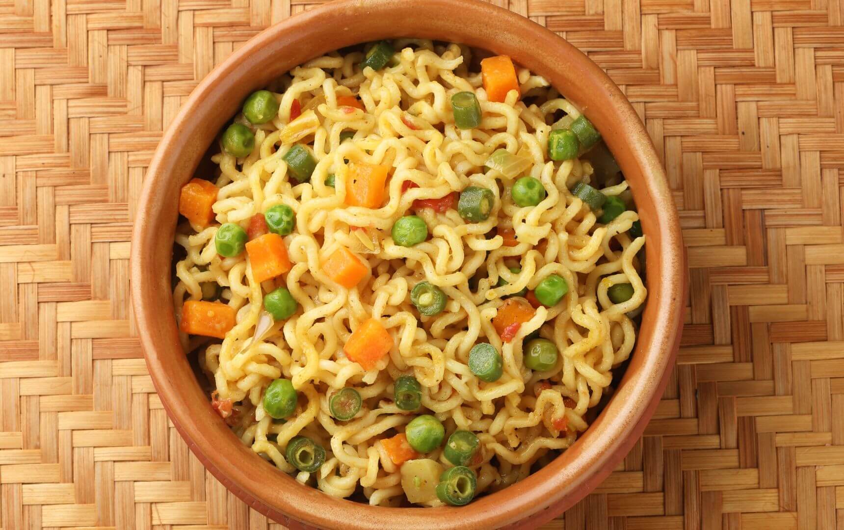 Bowl of nutritious noodles with peas and carrots, sitting on a woven placemat.