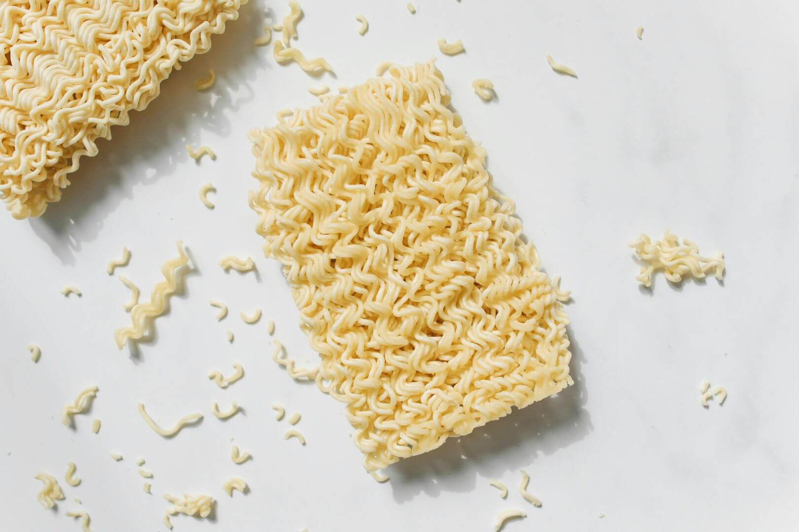 Blocks of instant noodles with some pieces broken off, all sitting on a white table background